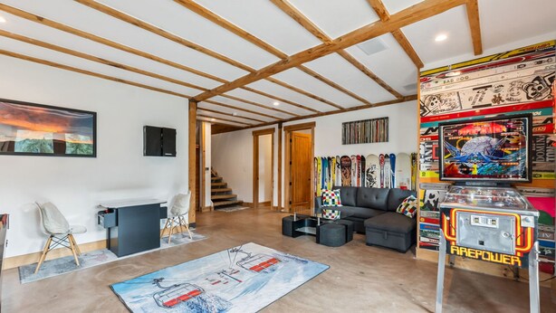 Ein modernes Wohnzimmer mit freiliegenden Holzbalken, einem Kamin, einer Couch, einem Spieltisch und einer Treppe, die in einen weiteren Raum führt.