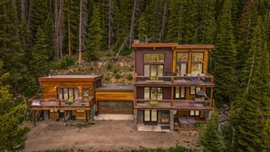 Eine moderne, mehrstöckige Hütte mit einer Terrasse und Sitzgelegenheit im Freien, umgeben von dichtem Wald.