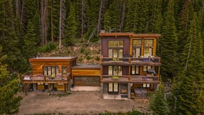 Eine moderne, mehrstöckige Hütte mit einer Terrasse und Sitzgelegenheit im Freien, umgeben von dichtem Wald.