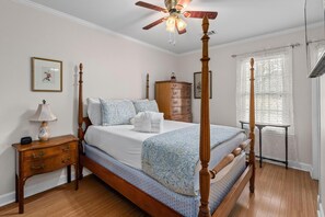 A four-poster bed with a wooden frame, a nightstand with drawers, a bedside table with a lamp, and a window with sheer curtains. - The Victorian Jewel Pet Friendly (Mount Pleasant)