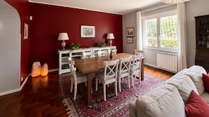 A dining area with a wooden table and chairs, a white cabinet with plants, and a large window with curtains.