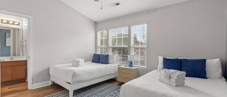 A bedroom with two beds, a ceiling fan, a window with blinds, and a bathroom with a mirror and sink.