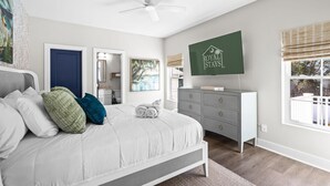 Un dormitorio con cama, cómoda, ventilador de techo, un cuadro y ventana con persianas.