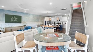Una sala moderna con un sofá, una mesa de centro de vidrio y un juego de Scrabble. Se ve una escalera que lleva a una zona de cocina con gabinetes blancos y una mesa de comedor.