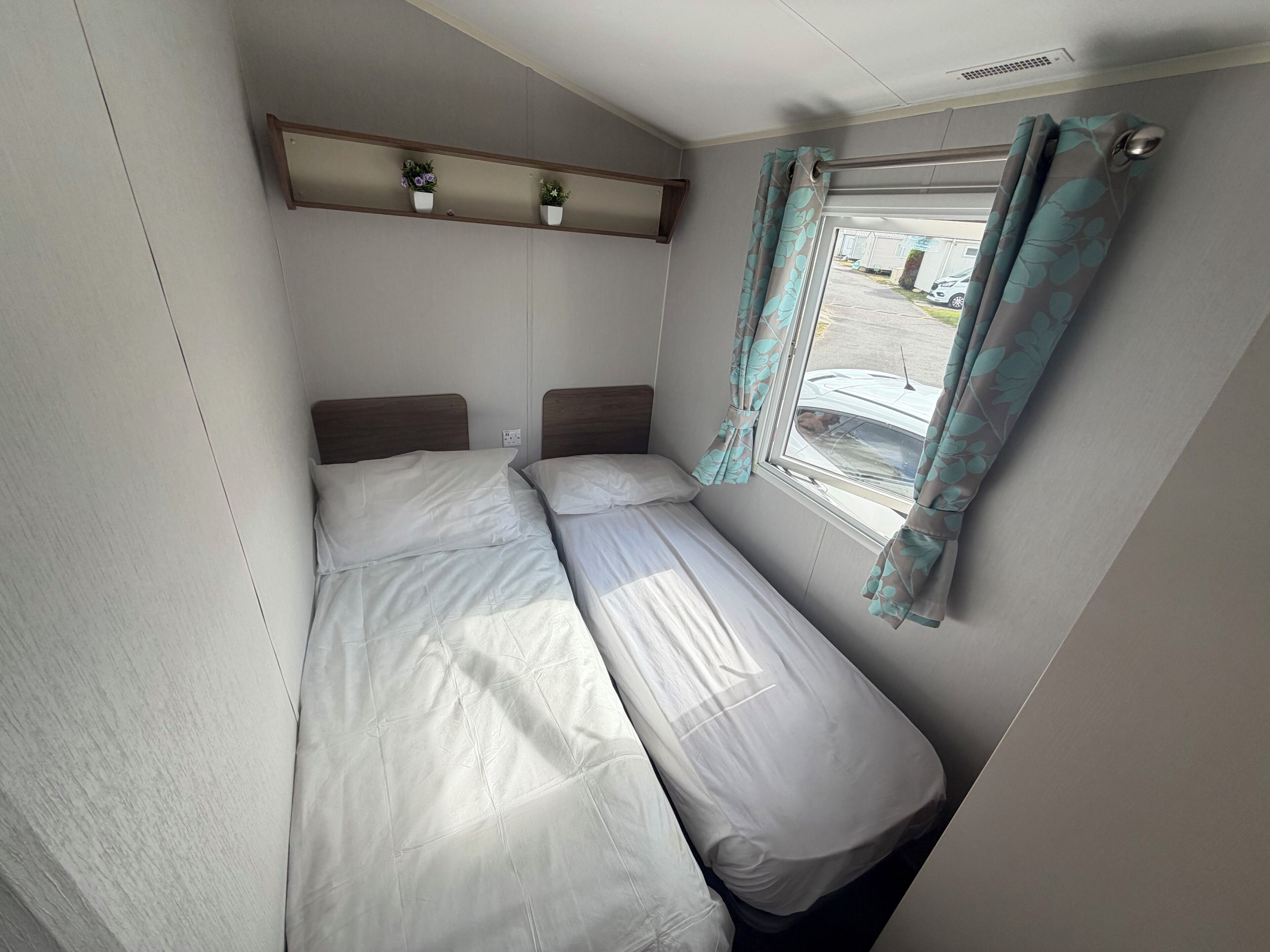 A small, single-bed room with a window, a shelf with potted plants, and a curtain.