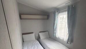 Una habitación compacta con dos camas, una ventana con cortinas y una estantería fijada a la pared.