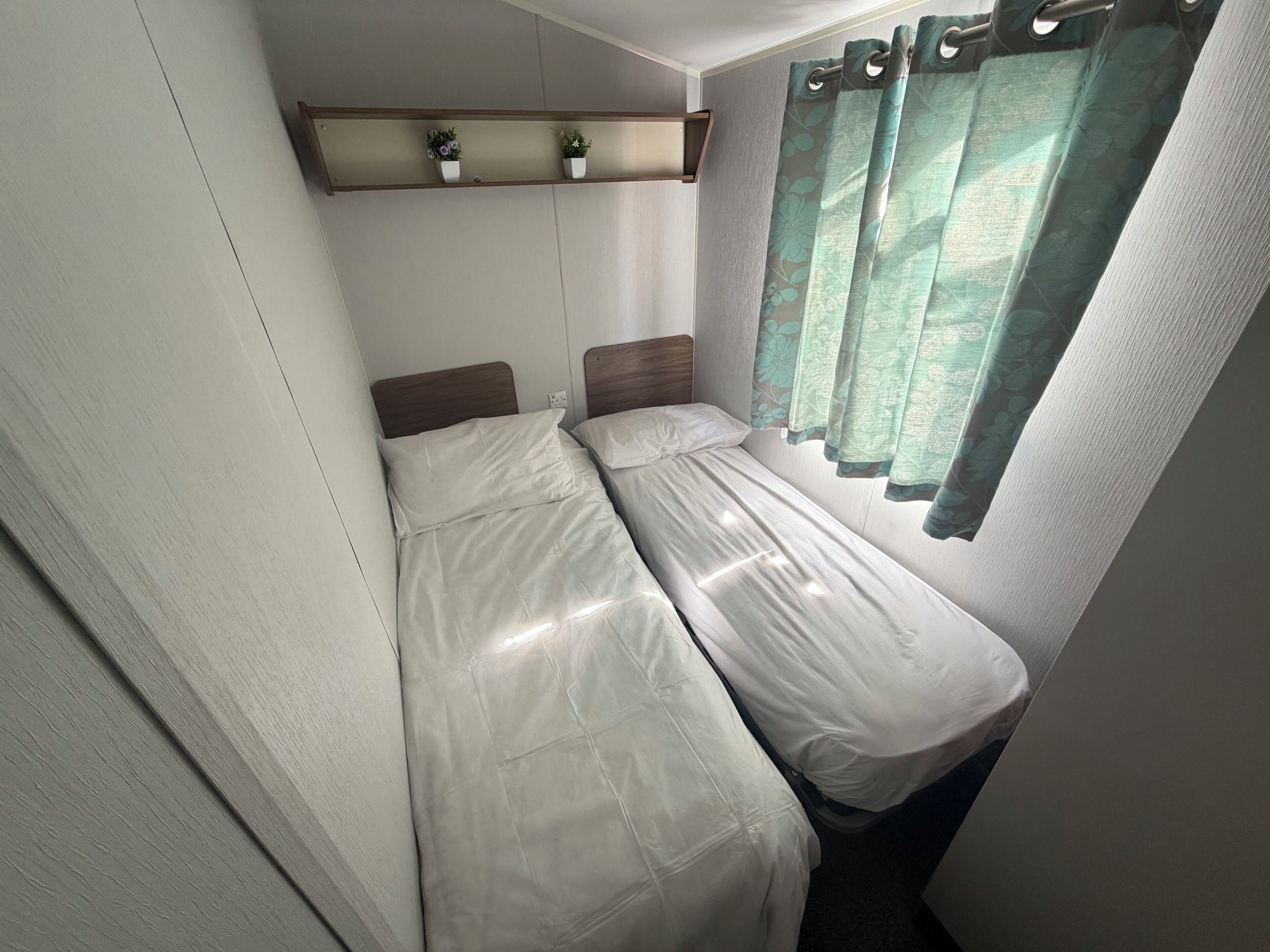 A compact room with two single beds, a shelf with potted plants, and a window with curtains.