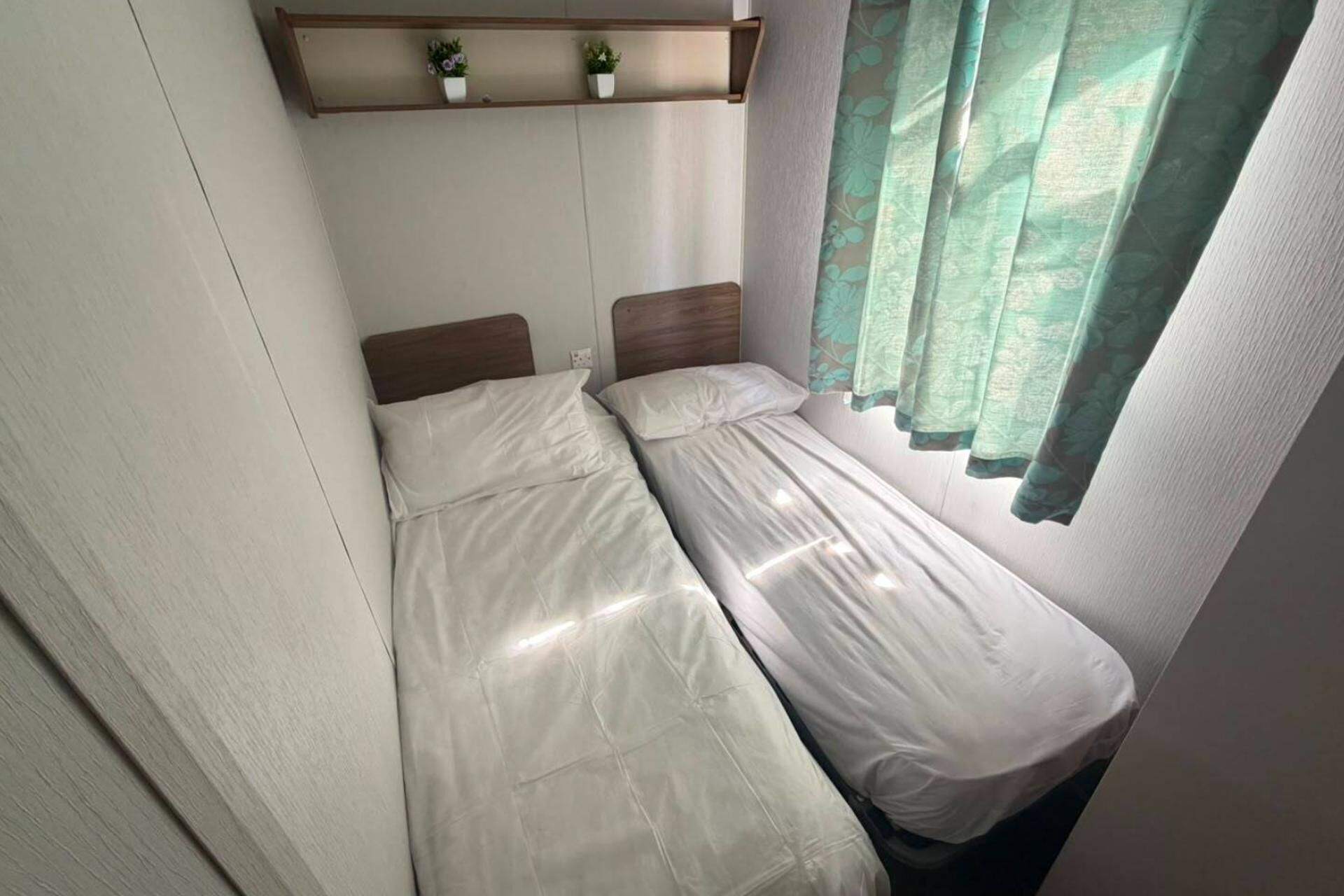 A small, single-bed room with a wooden headboard, a shelf with potted plants, and a window with a green curtain.