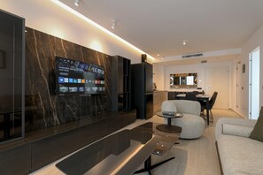 A modern living room with a large flat-screen TV, a sofa, a coffee table, and a dining area with a chair and table. - Tilosofi Luxury Apartments (Sarande)