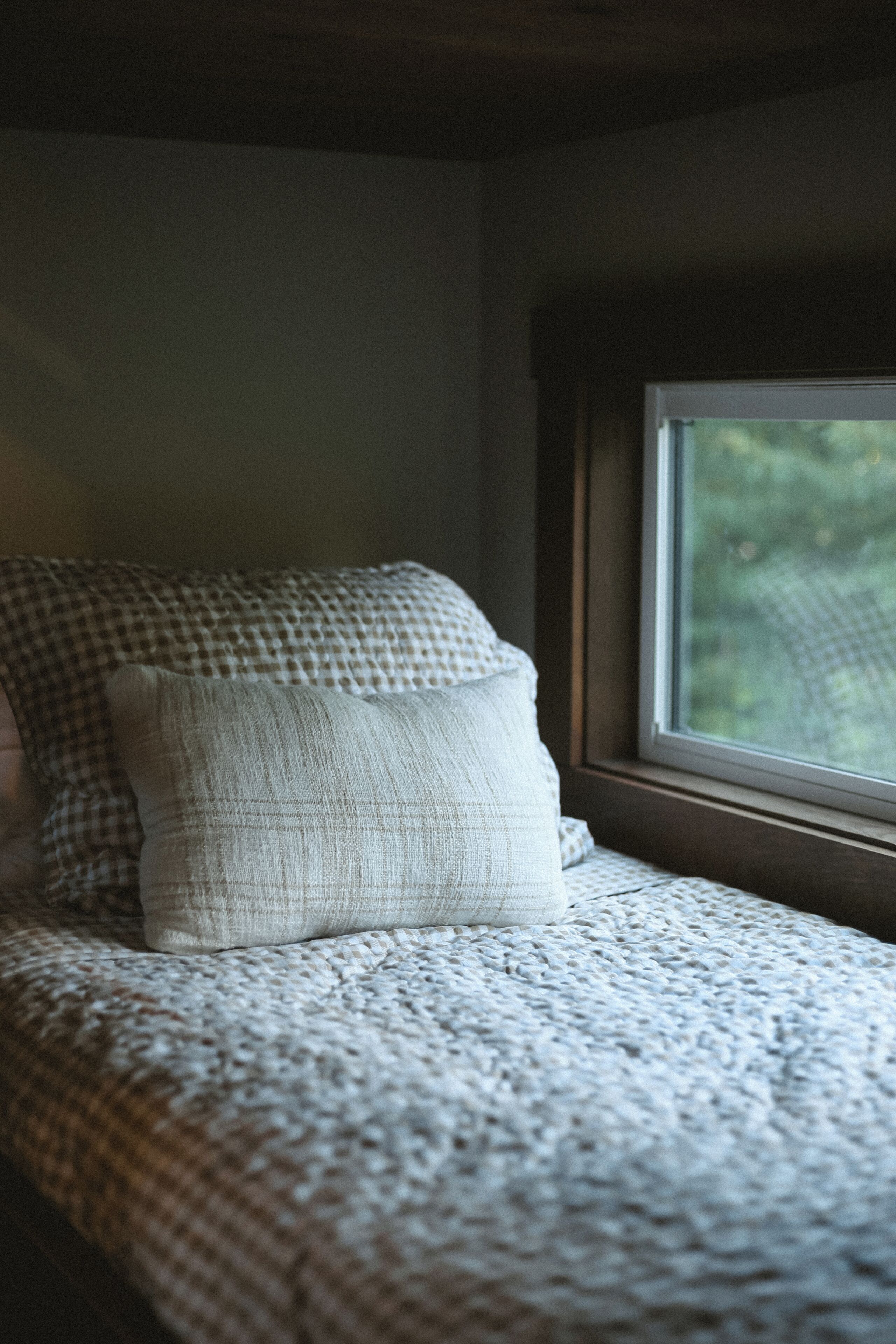 A bed with a knitted blanket and pillows, a window with a view of trees, and a wooden headboard.