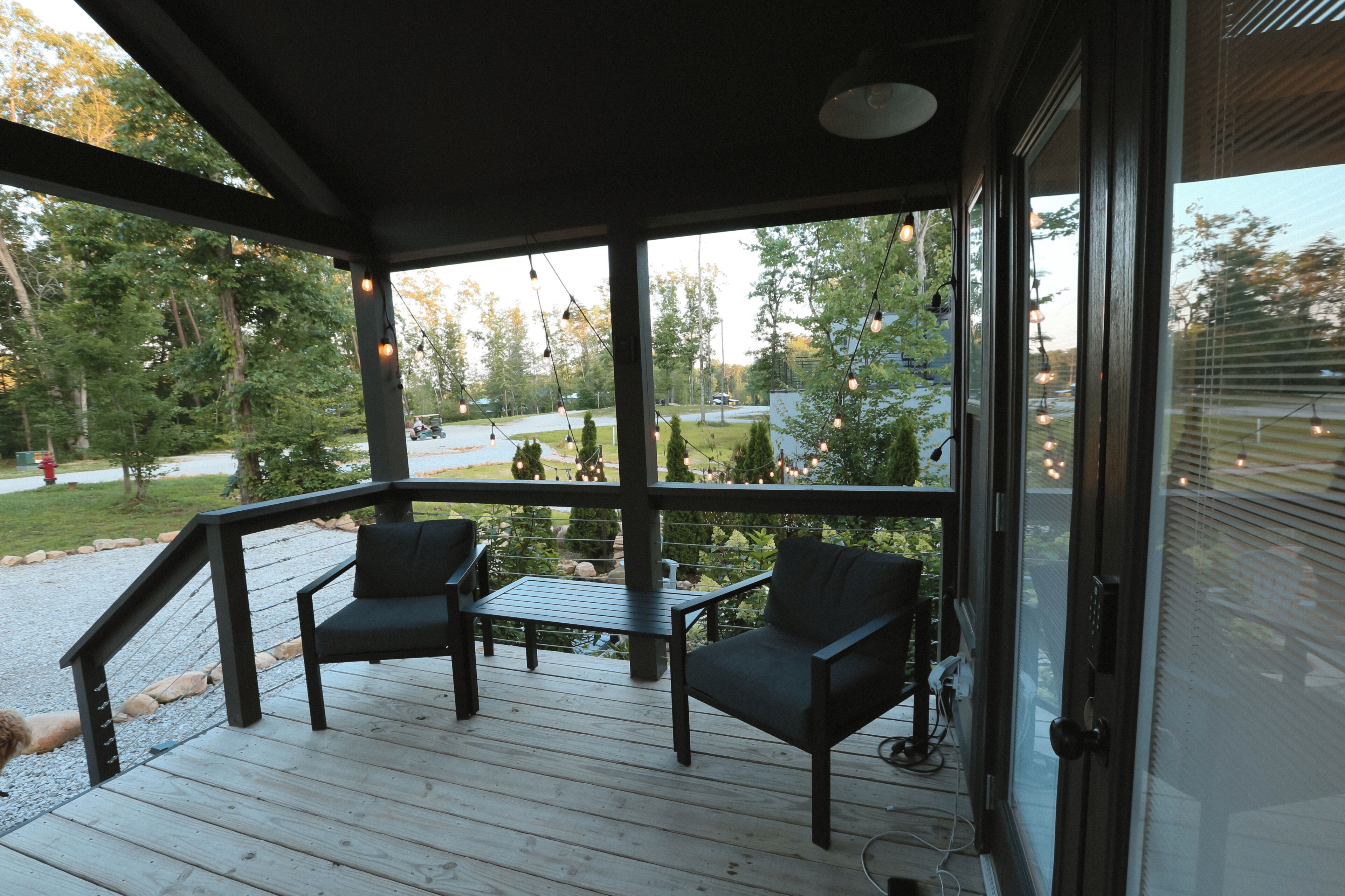 A covered outdoor seating area with two chairs and a table, overlooking a landscaped yard with trees and a distant view of a road.