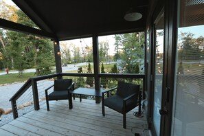 Un área de estar al aire libre con techo, con dos sillas y una mesa, con vistas a un jardín ajardinado con árboles y una vista distante de una carretera.