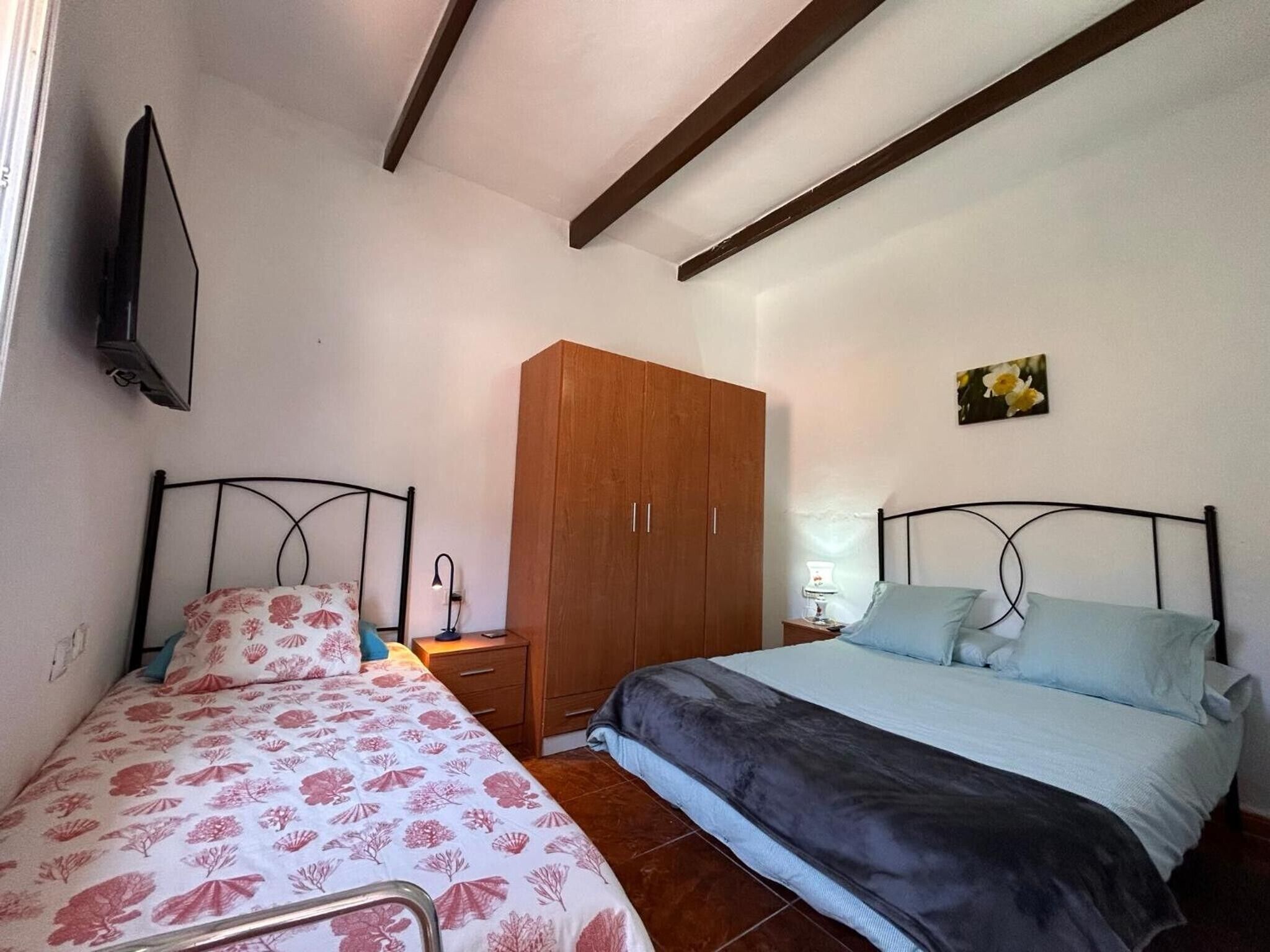Un dormitorio con dos camas, un armario de madera, un televisor montado en la pared y una mesita de noche con una lámpara.