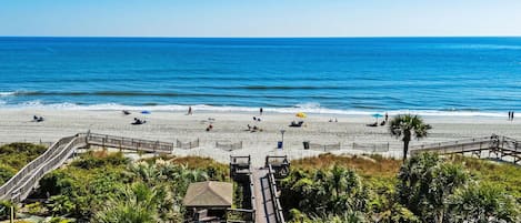 A beach with a wooden boardwalk leading to a gazebo, palm trees, and a clear blue sky.
