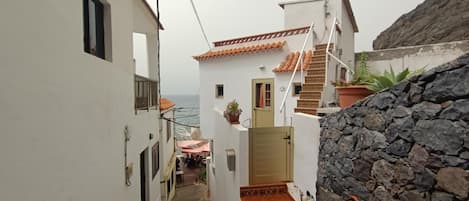 A narrow street with white buildings, a stone wall, and a staircase leading to a door.