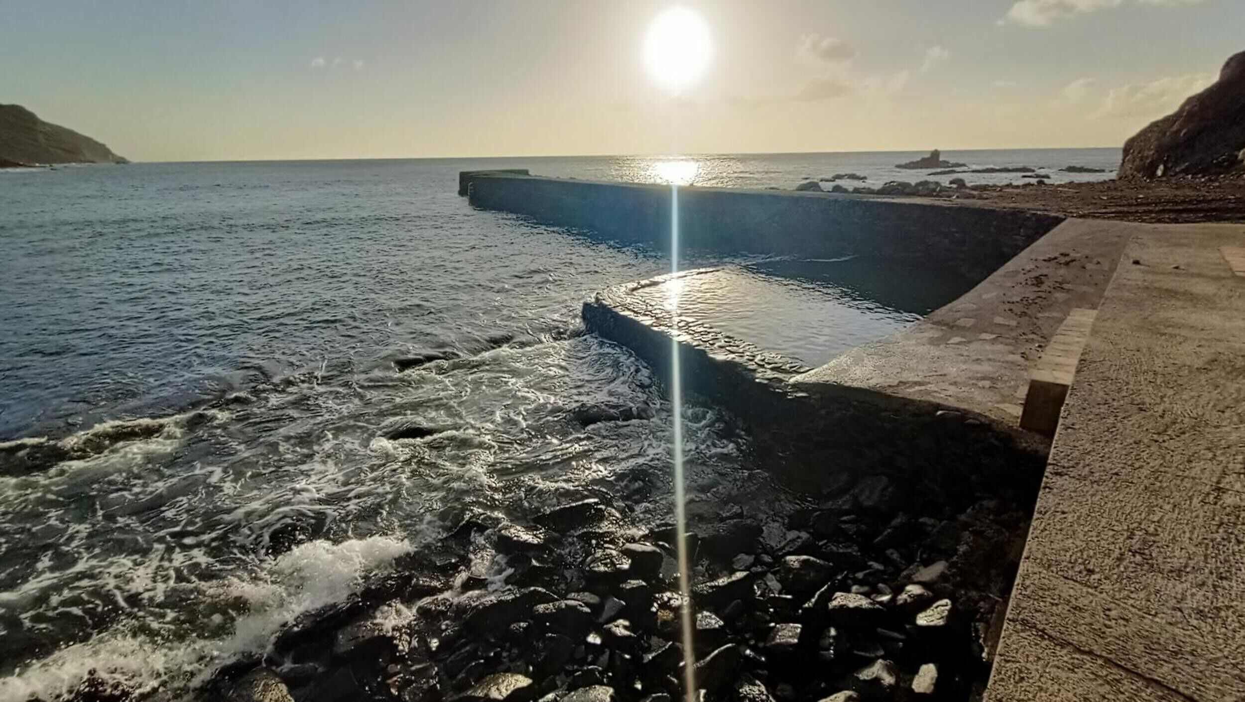 Een kustlandschap met golven die tegen een betonnen barrière slaan, een heldere lucht en een lage zon aan de horizon.