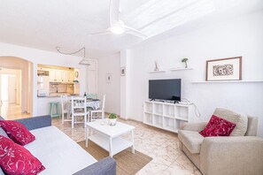 Ett vardagsrum med en soffa, ett soffbord, en tv, en takfläkt och ett kök i bakgrunden, samt en matplats.