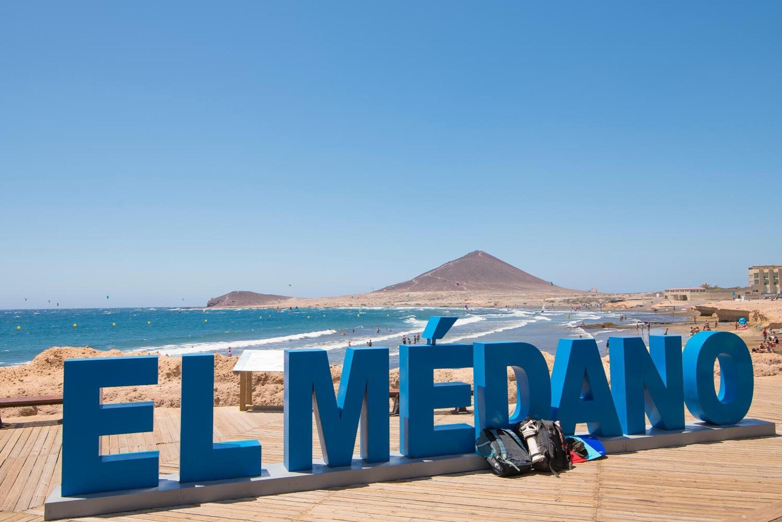 Eine Strandansicht mit dem Text 'EL MÉDANO' in großen blauen Buchstaben, einer Holzpromenade und einem Berg im Hintergrund.