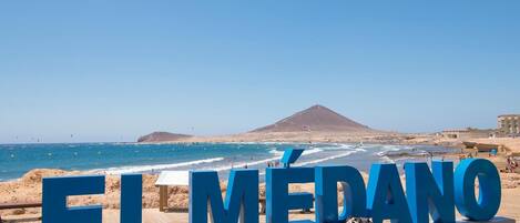 Eine Strandansicht mit dem Text 'EL MÉDANO' in großen blauen Buchstaben, einer Holzpromenade und einem Berg im Hintergrund.