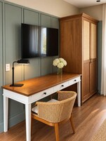 Una habitación con un escritorio de madera, un televisor montado en la pared, un armario y una silla de mimbre.