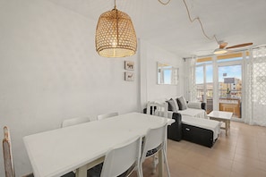A dining area with a white table and chairs, a woven light fixture, a sofa, and a view through a glass door. - Vacation Apartment 'Alquilaencanarias-medano Lagos De' with Mountain View, Pool & Wi-Fi (El Médano)