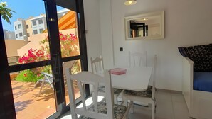 A dining area with a white table and chairs, a small white cabinet, and a view through a glass door to an outdoor patio with plants and a building. - Vacation Apartment 'Alquilaencanarias Duplex Beach' with Balcony and Wi-Fi (El Médano)