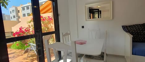 Un comedor con una mesa y sillas blancas, un pequeño armario blanco y vistas a un patio exterior con plantas y un edificio.