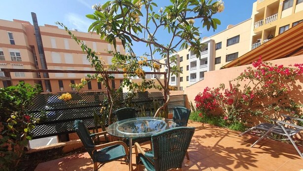 An outdoor patio with a round glass table and four chairs, surrounded by plants and a building in the background. - Vacation Apartment 'Alquilaencanarias Duplex Beach' with Balcony and Wi-Fi (El Médano)