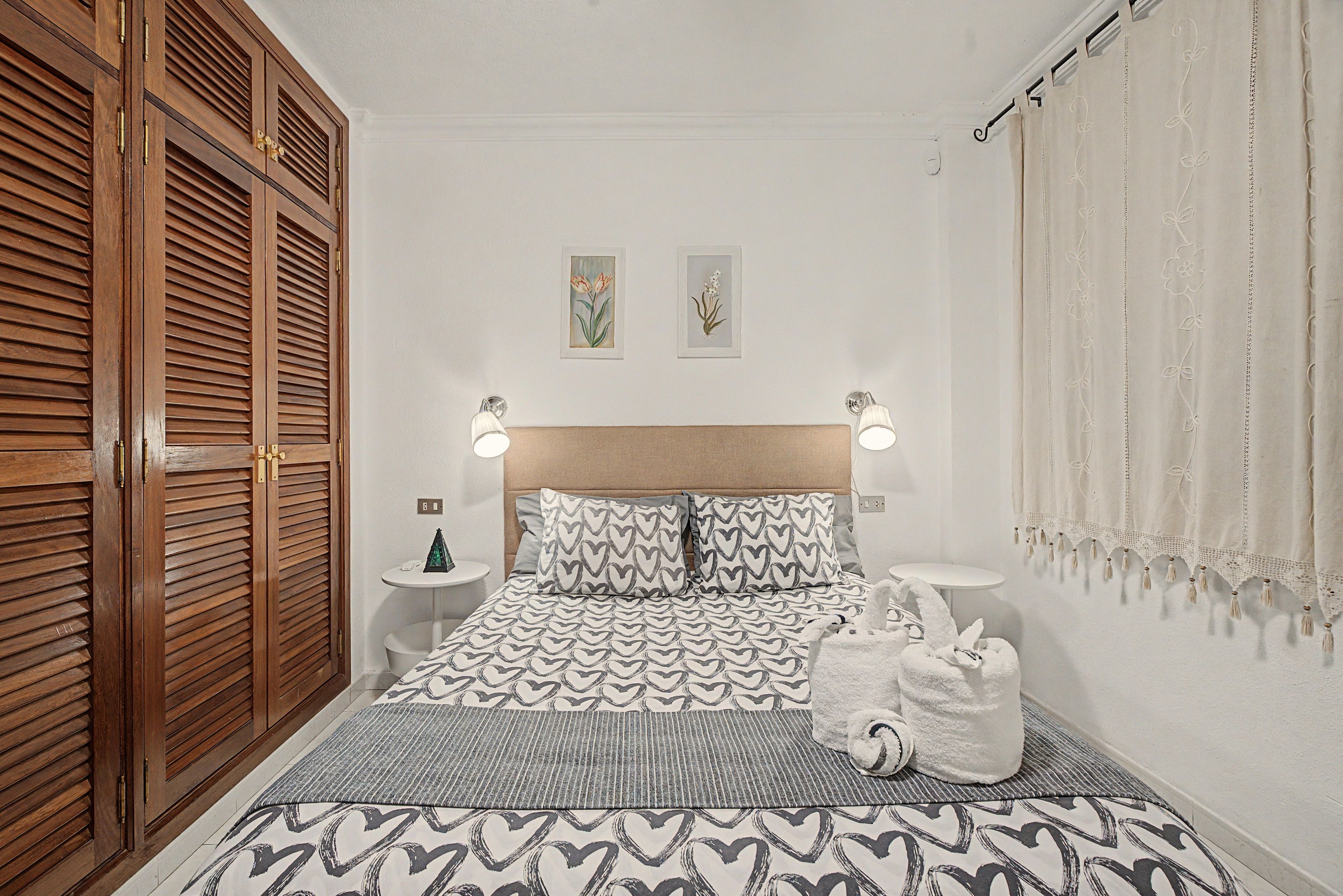 Une chambre avec une armoire en bois, un lit avec une literie à motifs, deux lampes fixées au mur et une petite table de chevet avec une lampe.