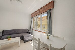A room with a grey sofa, a white table, and a window with curtains. - Vacation Apartment 'Alquilaencanarias-medano Tío' with Wi-Fi (El Médano)