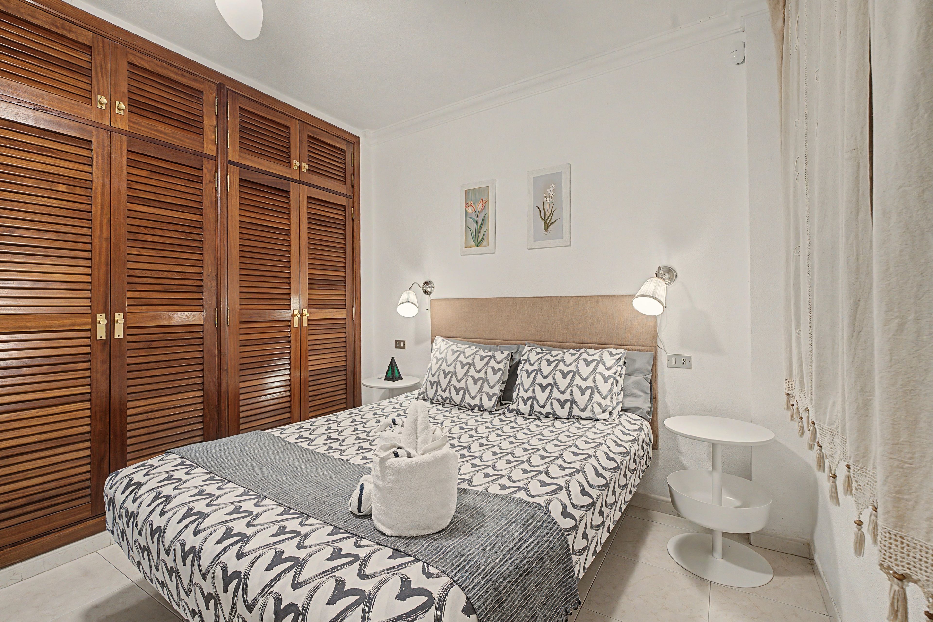 Un dormitorio con cama, mesitas de noche, armario y dos lámparas de pared.