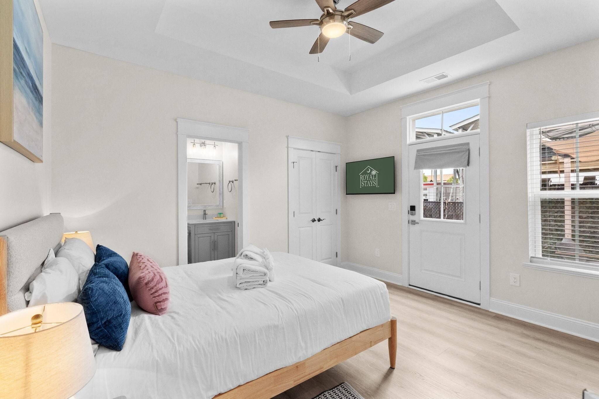 A bedroom with a bed, a ceiling fan, a bathroom with a mirror, a door, and a window with blinds.