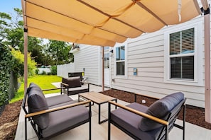 Een overdekt terras met buitenmeubilair, een barbecue en uitzicht op een woning.