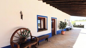 A covered outdoor area with a bench, a wooden wheel, a potted plant, and a wall with a door and windows.