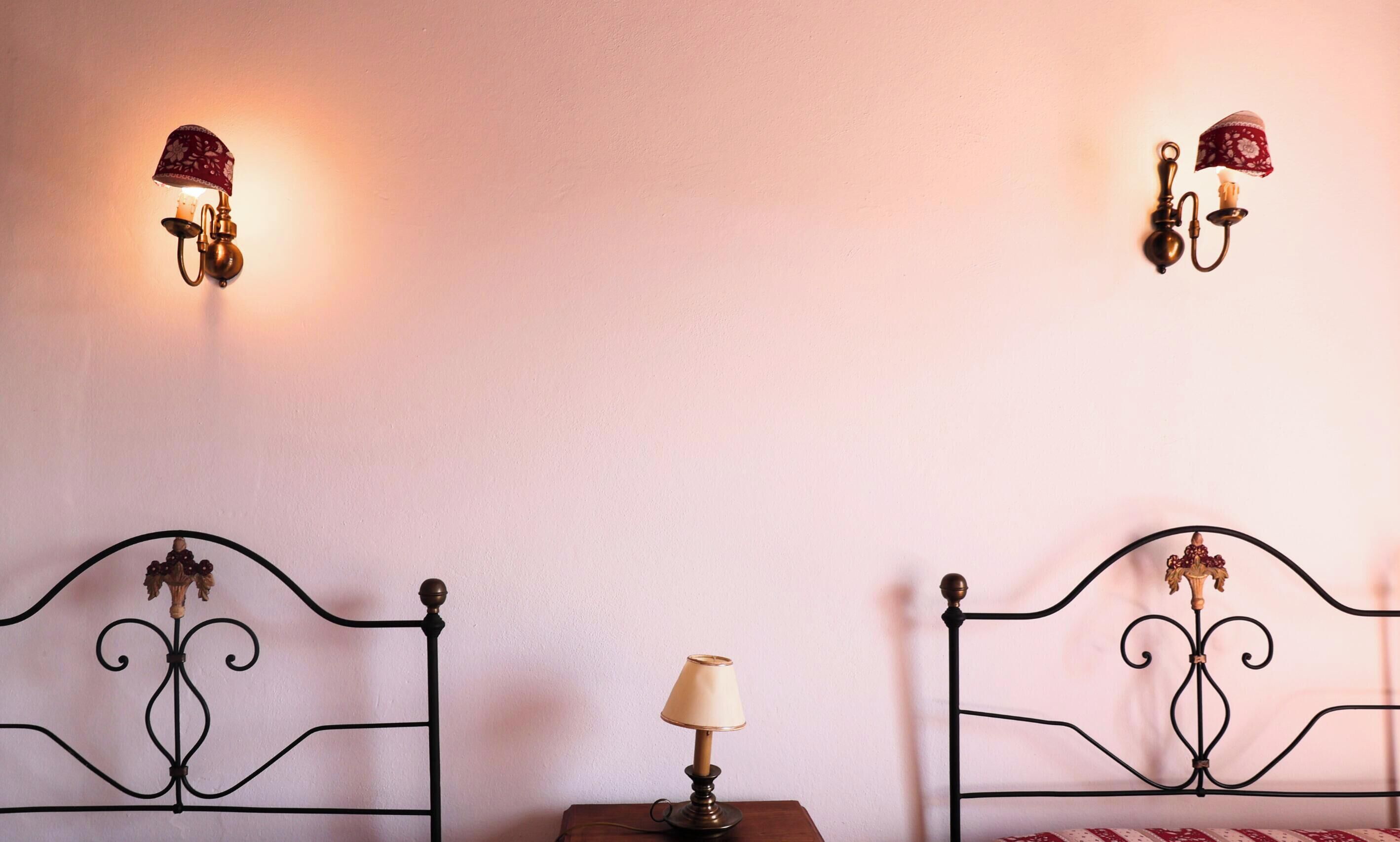 A room with two metal beds, a small table with a lamp, and two wall-mounted lamps.