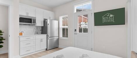 A modern hotel room with a kitchenette, a bed with white linens, and a view of the outside.