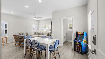 A modern kitchen with a white countertop, a dining area with a white table and blue and white patterned chairs, and a children's play area with a colorful arcade machine.