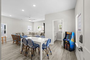 Una cocina moderna con encimera blanca, una zona de comedor con mesa blanca y sillas estampadas en azul y blanco, y un área de juegos infantiles con una máquina recreativa de colores.