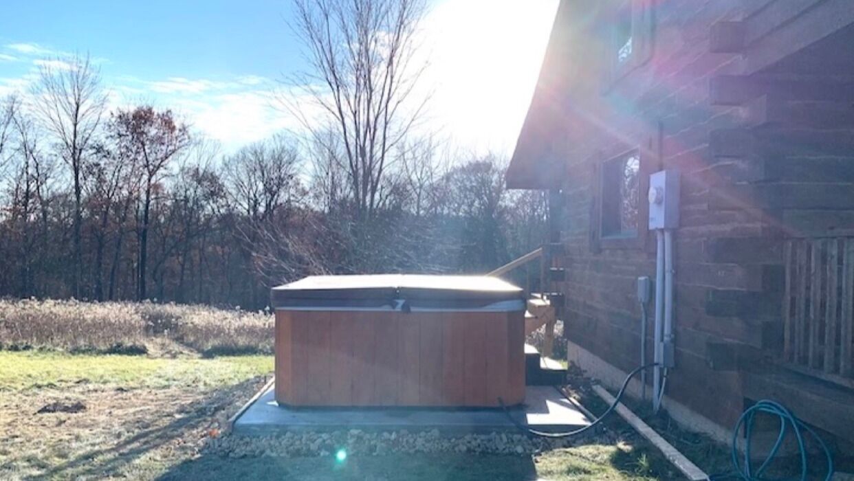 A hot tub outdoors next to a wooden cabin with a visible electrical outlet.
