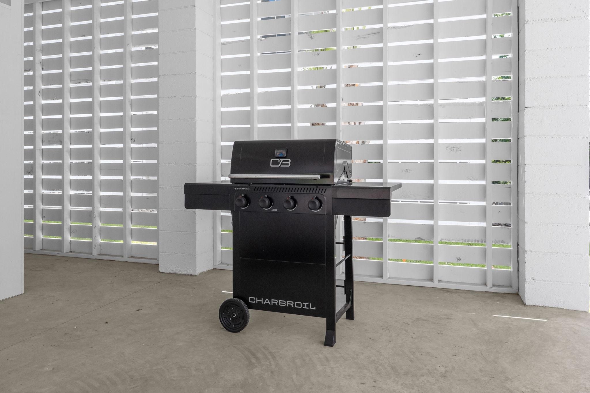 A black Charbroil grill with four burners and a side shelf, positioned in a room with white walls and a concrete floor.