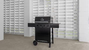 A black Charbroil grill with four burners and a side shelf, positioned in a room with white walls and a concrete floor.