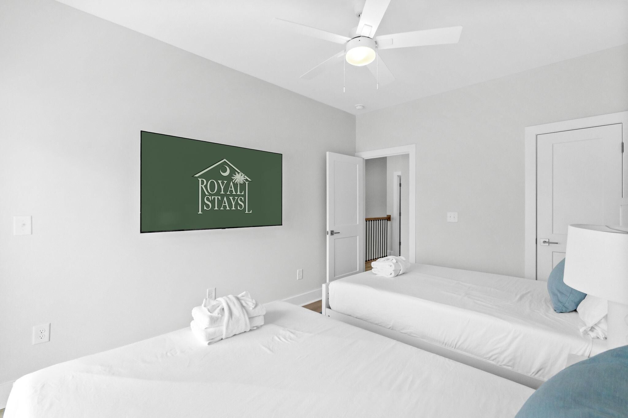 A hotel room with two beds, a ceiling fan, a sign reading 'ROYAL STAYS,' and a bathroom with a mirror and towel.
