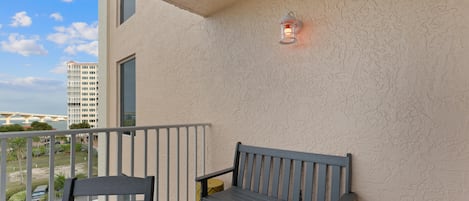 Un balcon avec un banc et une table, un tabouret jaune, un luminaire fixé au mur et une vue sur un immeuble et de la verdure.