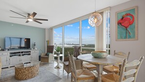 A spacious living area with a television, dining table, chairs, and a large window offering a view of the city. - Lovers Key #508 (Fort Myers Beach)
