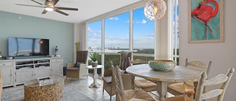 A spacious living area with a television, dining table, chairs, and a large window offering a view of the city.