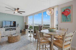A spacious living area with a television, dining table, chairs, and a large window offering a view of the city. - Lovers Key #508 (Fort Myers Beach)