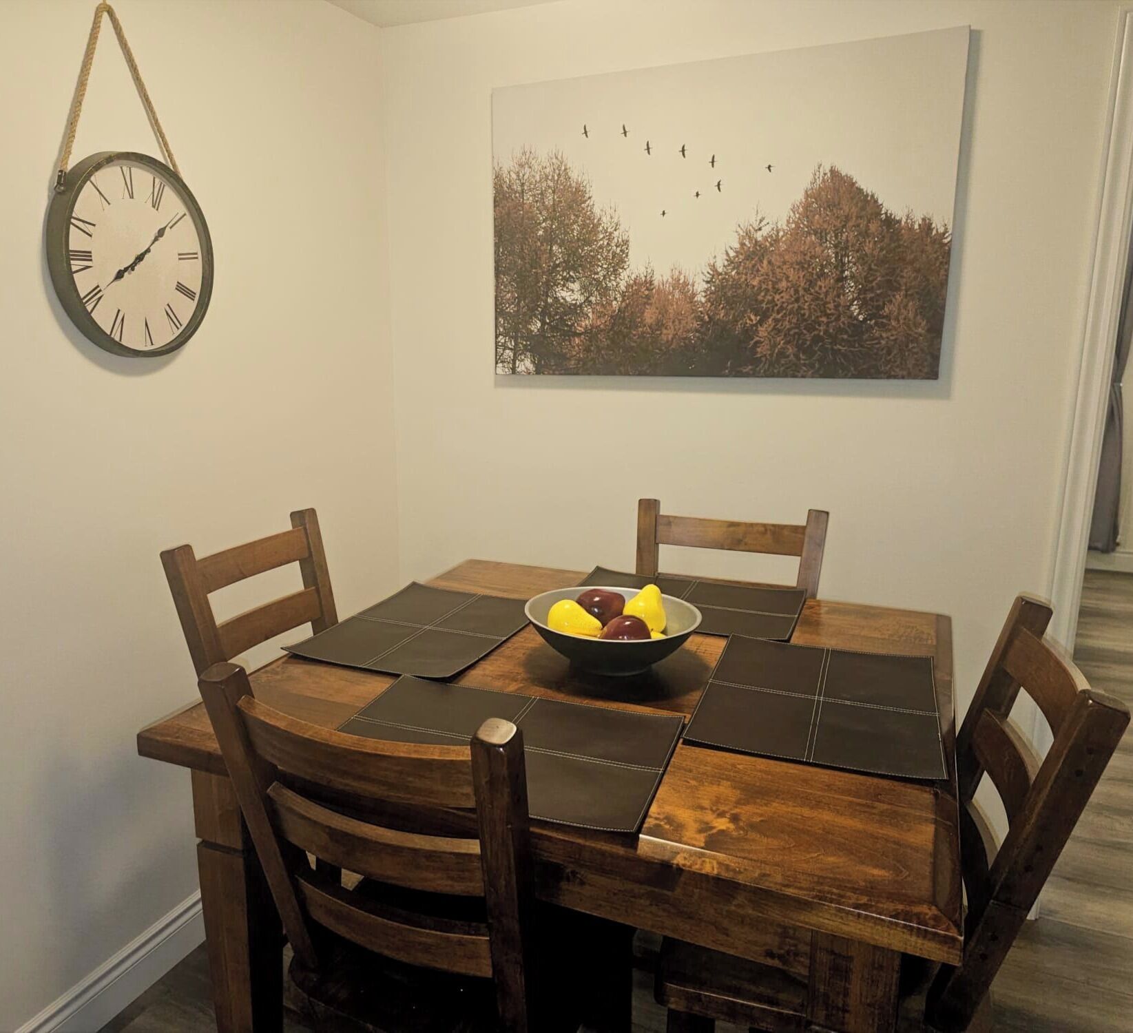 Una mesa de comedor con cuatro sillas, un frutero, un reloj de pared y un cuadro enmarcado.