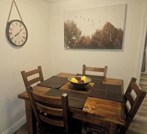 Une table à manger avec quatre chaises, un saladier rempli de fruits, une horloge murale et un tableau encadré.