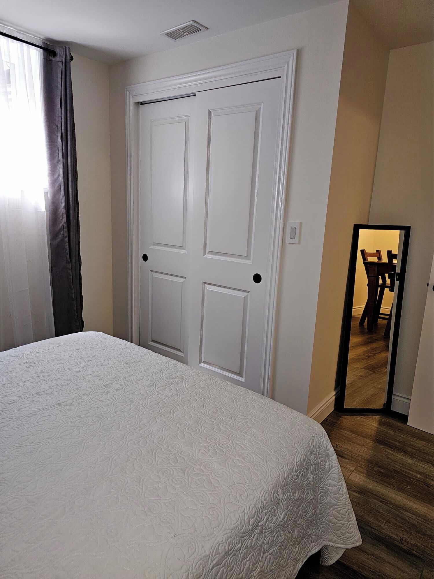 A room with a bed, a white door, and a mirror reflecting a dining area.