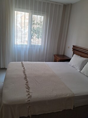 Un dormitorio con una cama grande, una mesita de noche y una ventana con cortinas traslúcidas.